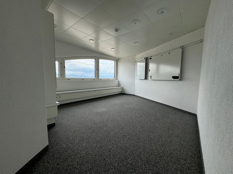 The image shows a spacious office space with a suspended ceiling, recessed lighting, and a large window overlooking the exterior. The room has gray carpeted flooring and white walls. There is a built-in bench or seating area along one wall, and a wall-mou