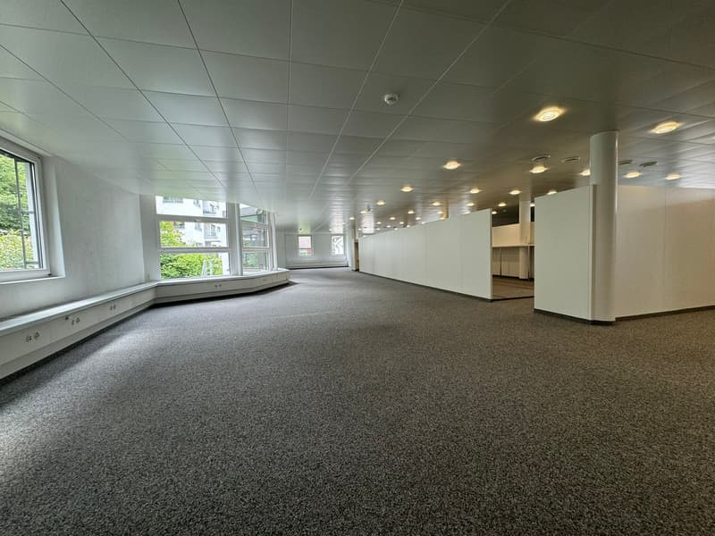 Large open office space with gray carpeted floor, white walls, and recessed lighting. The space has several windows providing natural light and appears to be a commercial or office setting.