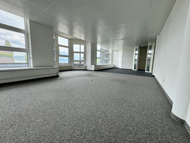 Large open office space with gray carpeted flooring, white walls, and large windows providing natural light. The space appears to be empty and ready for use.