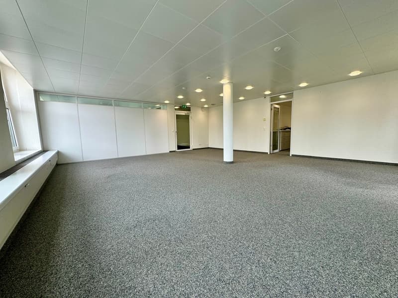 Large open office space with gray carpeted flooring, white walls, recessed lighting, and glass partitions. The space appears to be a commercial or office environment.