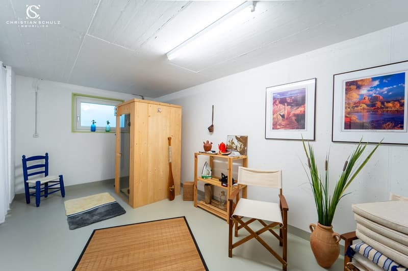 small room with white walls, wooden cabinet, wooden chair, small rug, wooden floor, wooden shelf, potted plant, framed pictures, small window