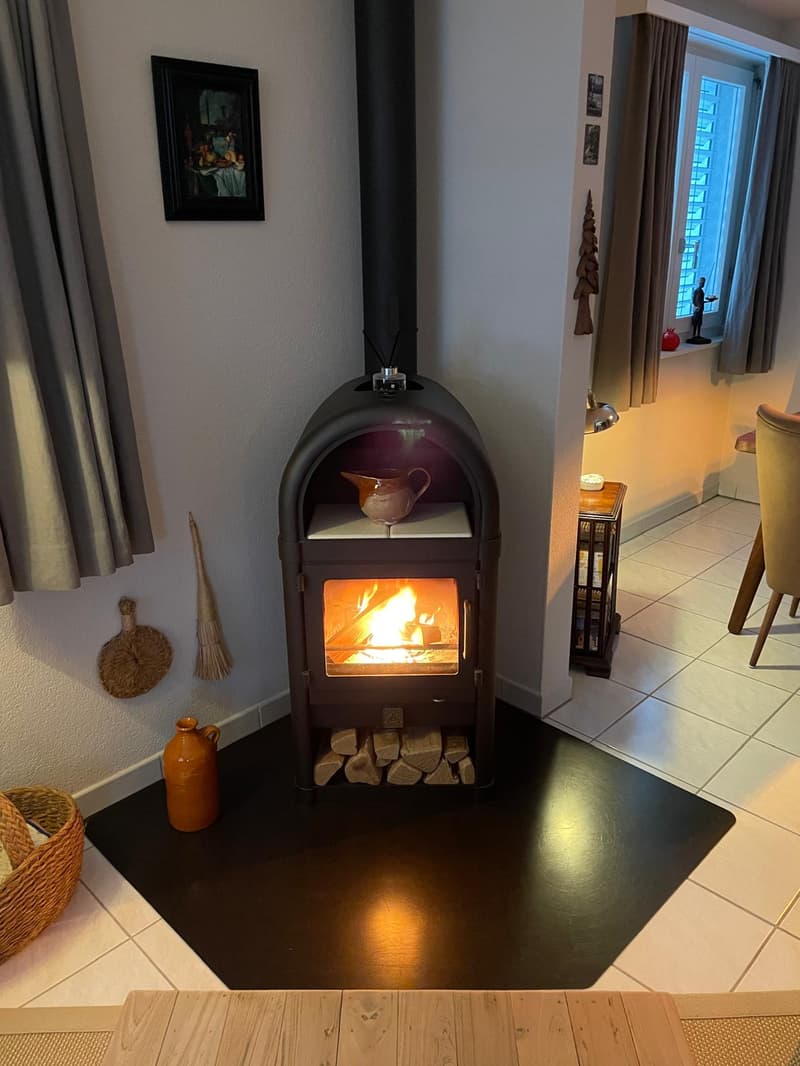 Fireplace in the corner, black mat, white tiles, wooden table, basket, picture frame, broom, jug