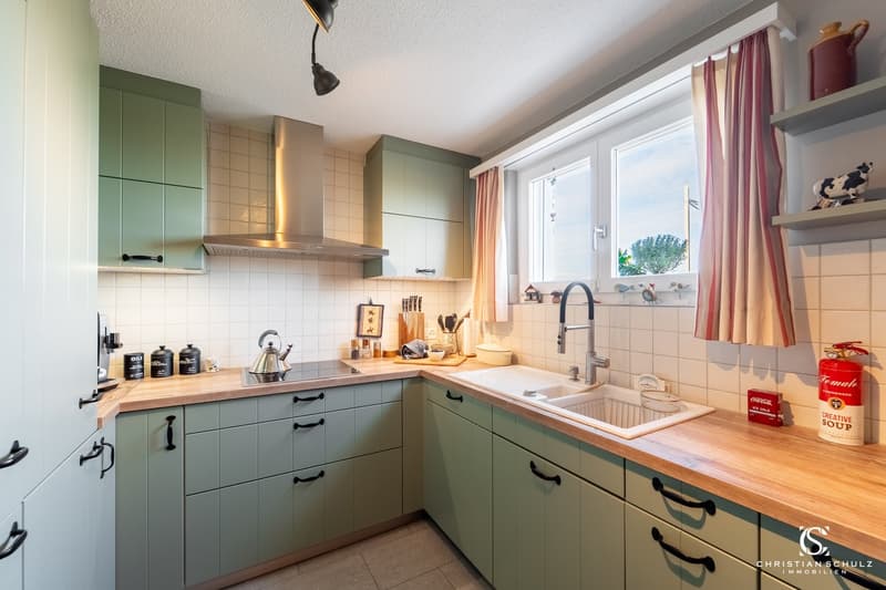 modern kitchen with wooden countertops, green cabinets, white tiled walls, stainless steel appliances, window with curtains, sink with faucet, fire extinguisher, Coca-Cola can, teapot, chopping board, knives, and various kitchen utensils