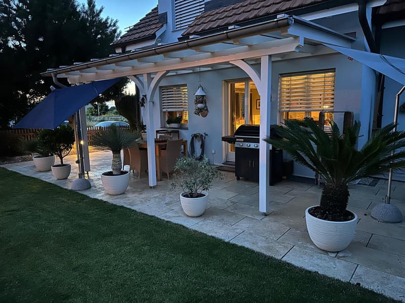 Patio with stone flooring, white pergola, dining table and chairs, barbecue, potted plants, blue umbrella, green lawn, house in the background