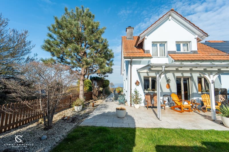 white house, brown roof, solar panels, wooden fence, patio, wooden chairs, potted plants, trees, grassy area