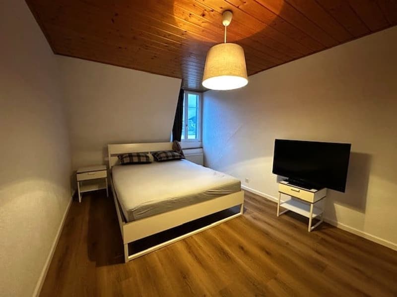 a white bed, pillows, wooden floor, TV on stand, window, lamp
