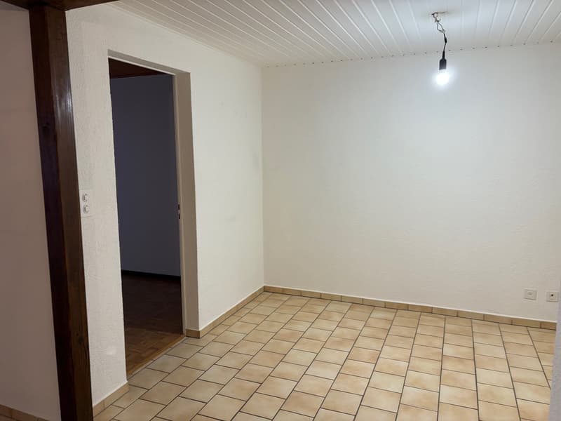 empty room with white walls, tiled floor, doorway to another room, open doorway to a hallway, single light fixture on the ceiling, light switch on the wall, power outlet on the wall