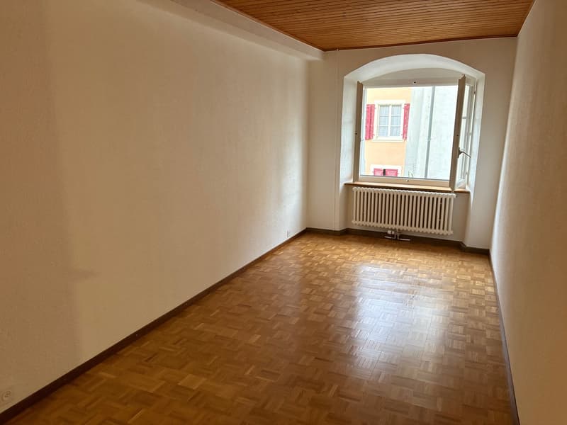 An empty room with wooden parquet flooring, white walls, a radiator beneath an arched window, and exposed wooden ceiling.