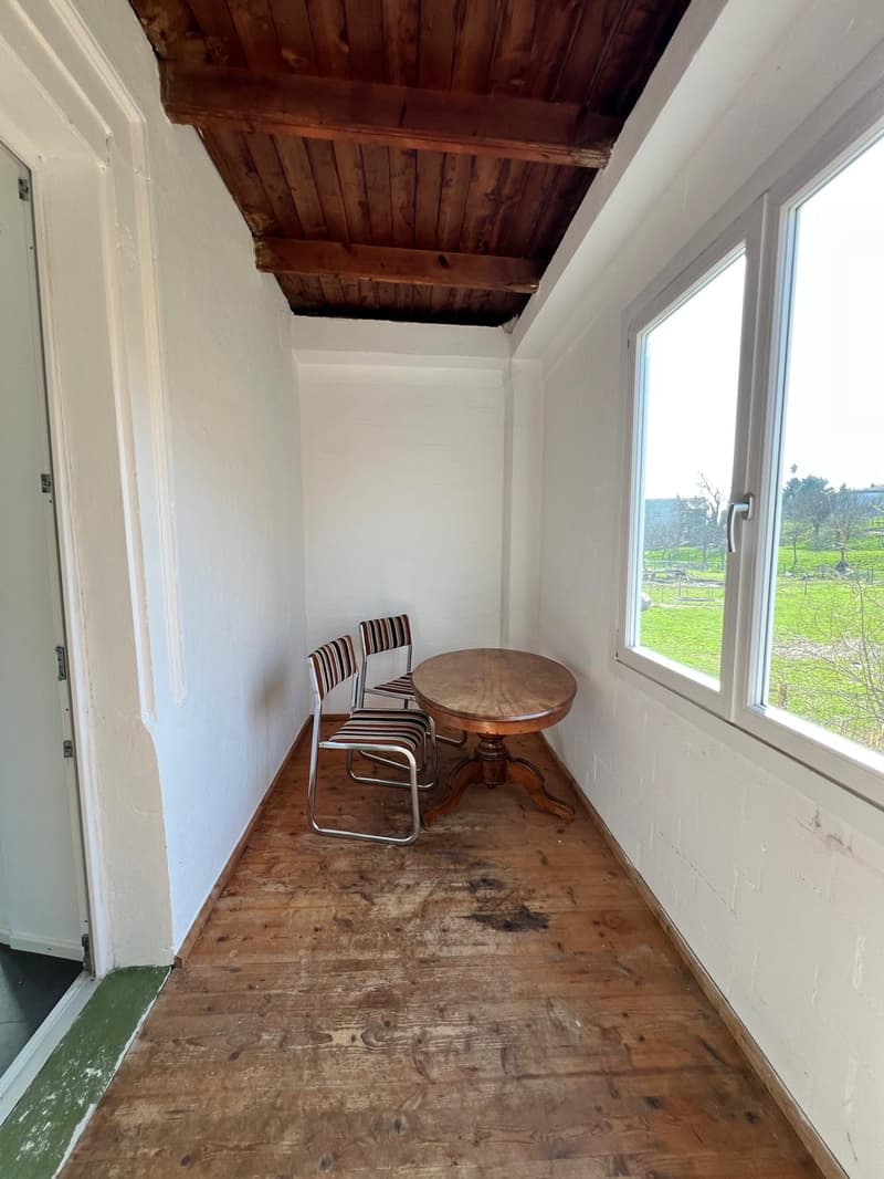 A small room with wooden floor, white walls, wooden ceiling, a round table, and two chairs. The room has a glass window showing a grassy field view.