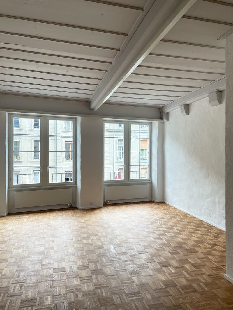 An empty room with two large windows, white walls, and wooden floors. There is a radiator on each wall next to the windows.