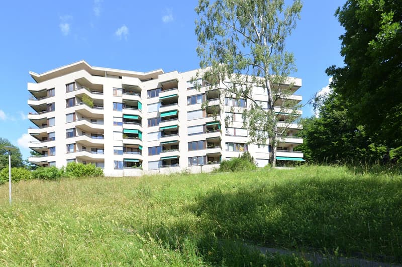 Modern white apartment building with multiple balconies, situated on a grassy hill