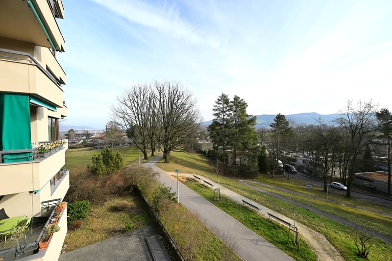 A building with balconies, a pathway, benches, trees, and a view of the city and mountains.