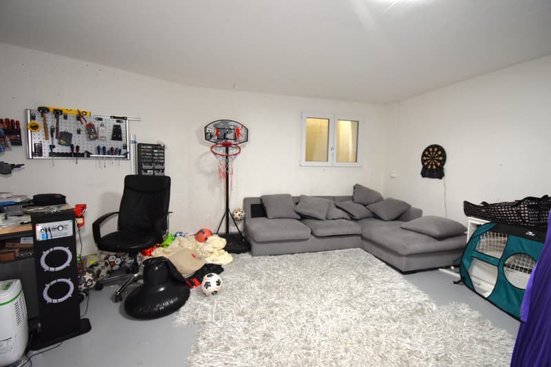 A room with a white rug, a gray sofa, a black chair, a basketball hoop, a dartboard, a speaker, and a desk with various items on it.