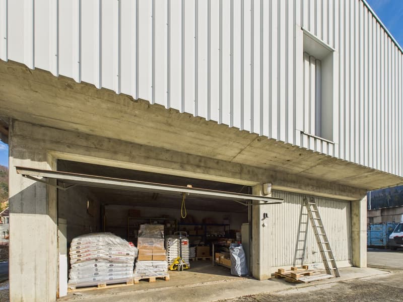 Industrial building with a concrete floor, white metal sheet wall, small window, a wide open garage door with a stack of wrapped materials inside