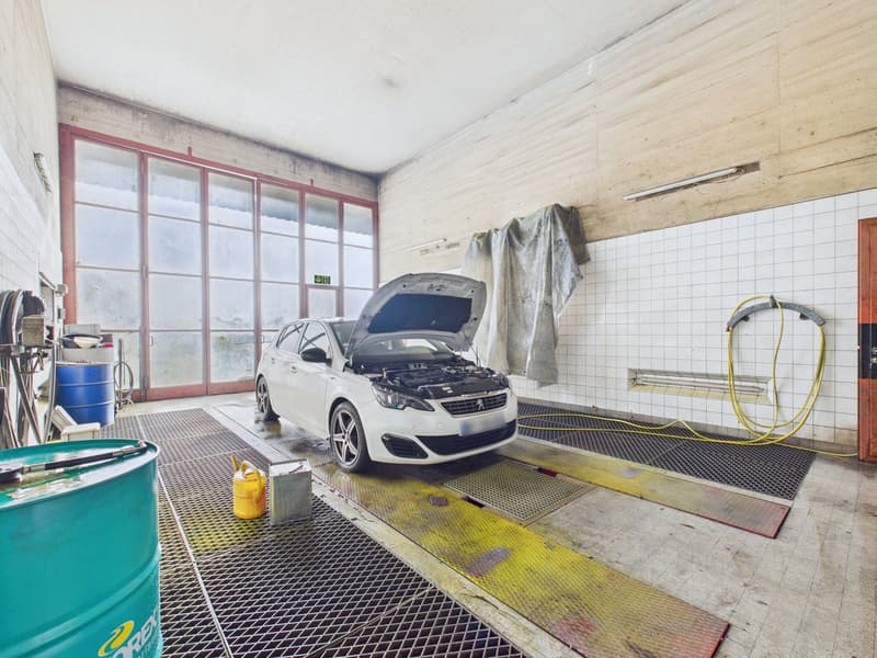 An enclosed car wash with large windows, white tile walls, drain on the floor, a white car with its hood up, oil can and drum nearby.