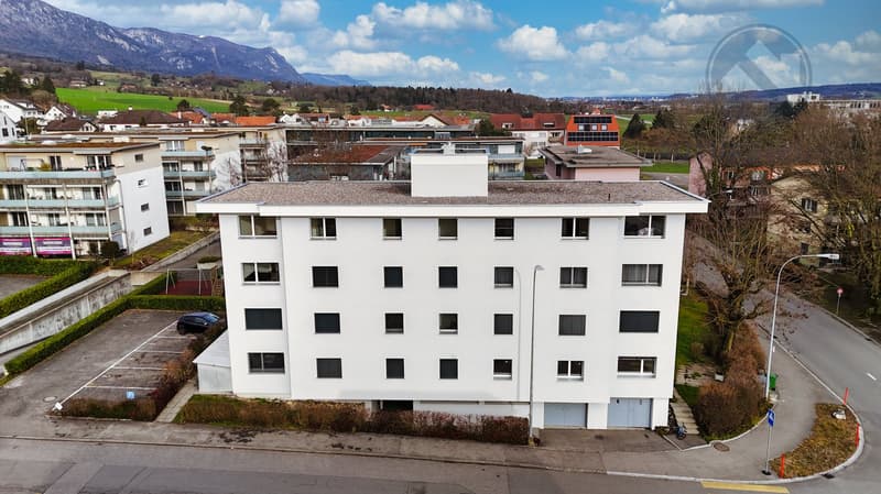 A modern white building with multiple windows, a flat roof, and a parking lot in front.