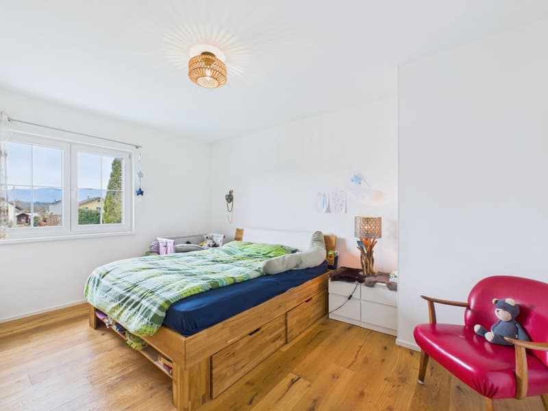 Bedroom with wooden bed, white walls, wooden floor, large window, bedside table, lamp, chair, stuffed toy, storage drawers under the bed