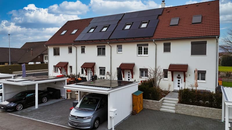 Two-story house with solar panels on the roof, white exterior, red roof, two garages, black car, gray van, brick driveway, small garden, chimney