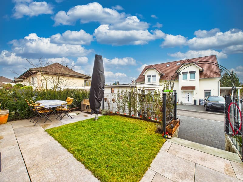 A garden with a table and chairs, a grassy area, and a car parked on the side of the road.