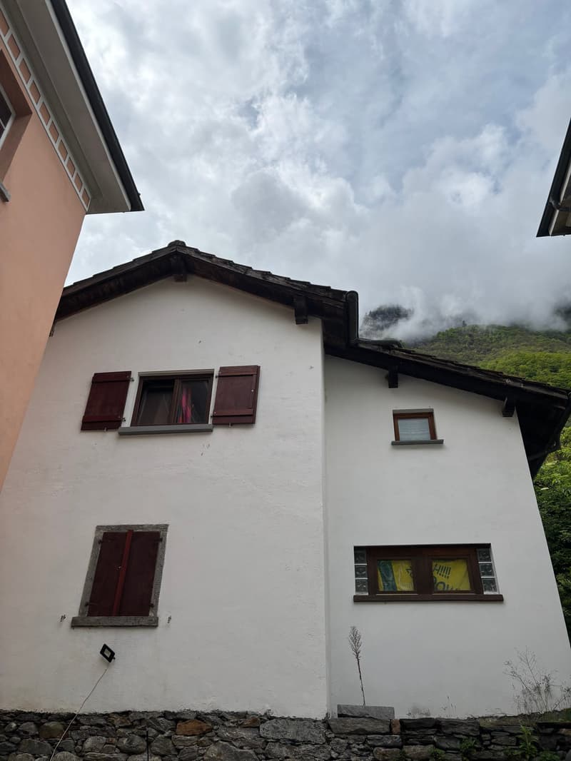 2-story white building with red-tiled roof, wooden shutters on the windows, and a small balcony or terrace visible. The building is set against a backdrop of cloudy skies and lush greenery.