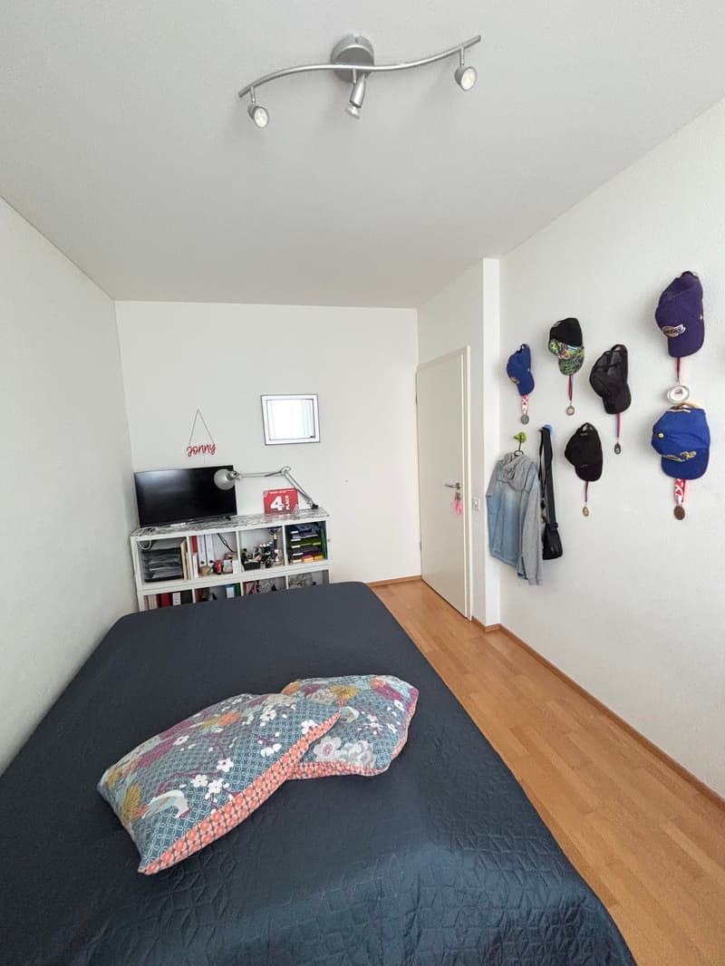Bedroom with a black bed, a TV stand with shelves, and a wall-mounted coat rack with various hats and jackets.