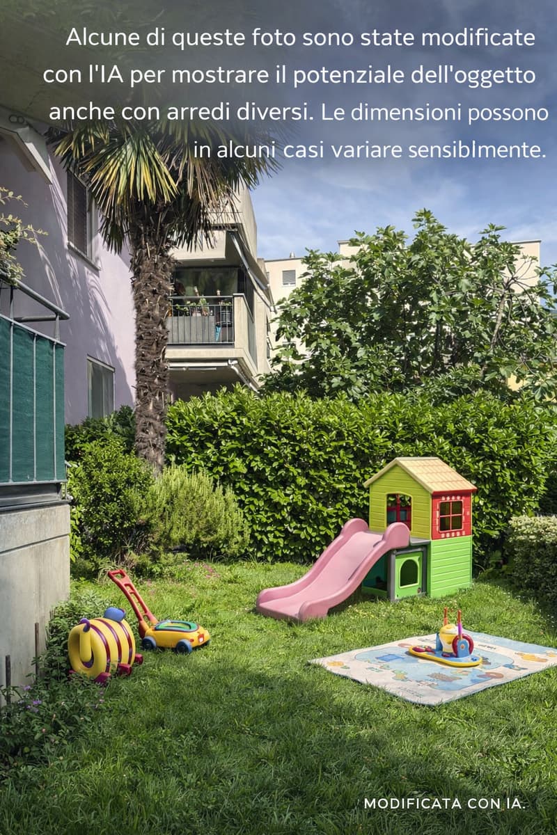 A garden with a slide, a playhouse, a toy car, a toy lawn mower, and a mat on the grass. There is a building with a balcony and a tree in the background.