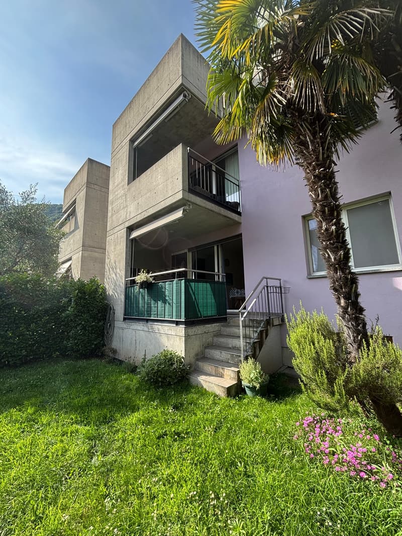 2-story concrete building with balconies, surrounded by a lush green lawn, palm trees, and other vegetation