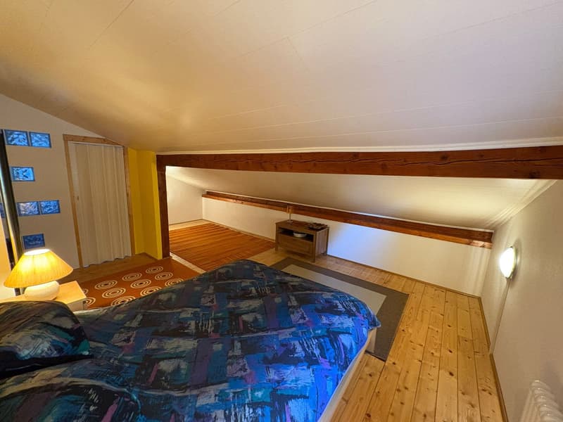 Small bedroom with wooden beams, wooden floor, double bed, lamps, small wooden side table, wooden rug, radiator, two rugs on the floor