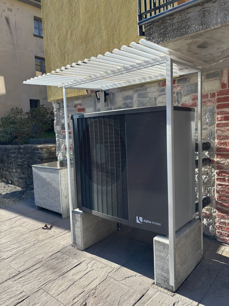 A grey outdoor electrical unit with a metal cover, mounted on concrete blocks, and a small desk with a faucet is visible in the image. The unit is probably a part of a larger building, and a staircase is visible on the upper left side.