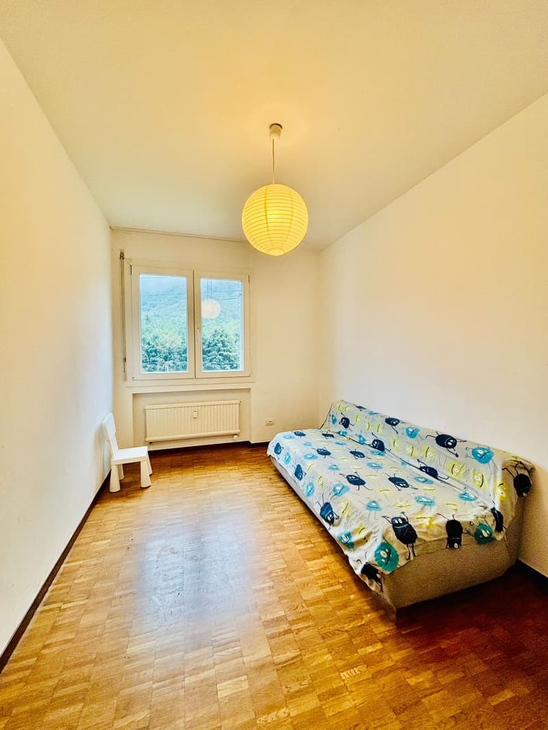 Room with double bed, white walls, wooden floor, a window with mountain view, white chair, and a hanging lamp