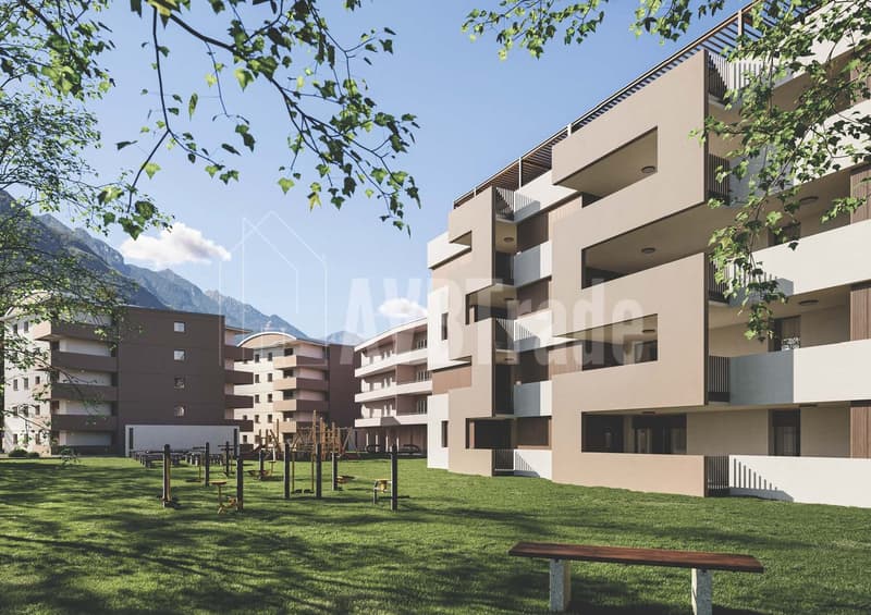 Multi-story apartment building with balconies, surrounded by a grassy area with playground equipment and benches, with mountains visible in the background.