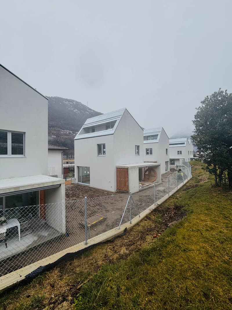White modern houses in a row, under construction, surrounded by grass and a fence, with a mountain in the background.