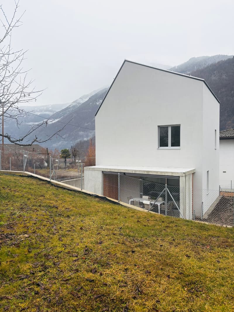 A single-story, white exterior house, elevated on the slope, featuring a garage with roll-up doors and metal fencing for perimeter security, set against a backdrop of mountainous scenery.