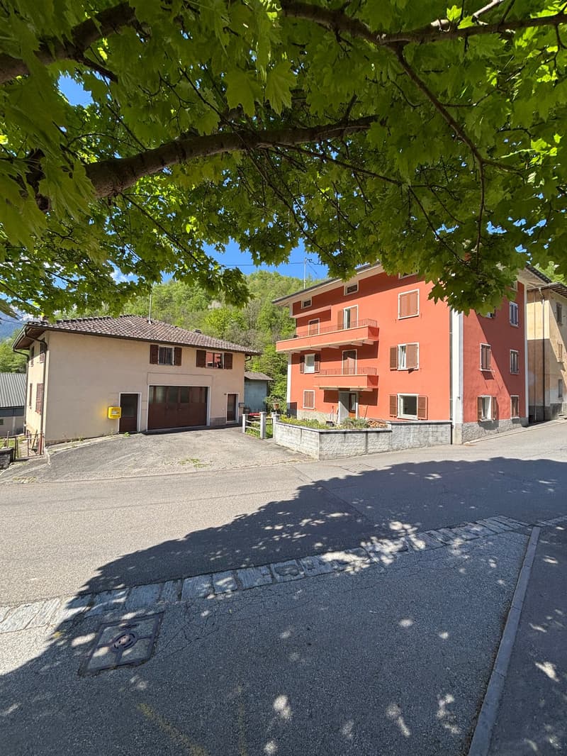 The image shows a residential area with several multi-story apartment buildings. The buildings have a reddish-orange exterior color and balconies. There are trees with lush green foliage overhanging the scene, creating a shaded and natural environment. Th