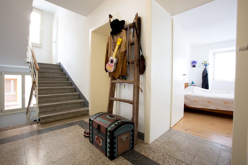 The image shows the interior of a home, with a staircase leading up to the second floor. There is a wooden ladder-like structure on the wall, with various coats and accessories hanging from it. A large suitcase or trunk is visible on the floor near the st