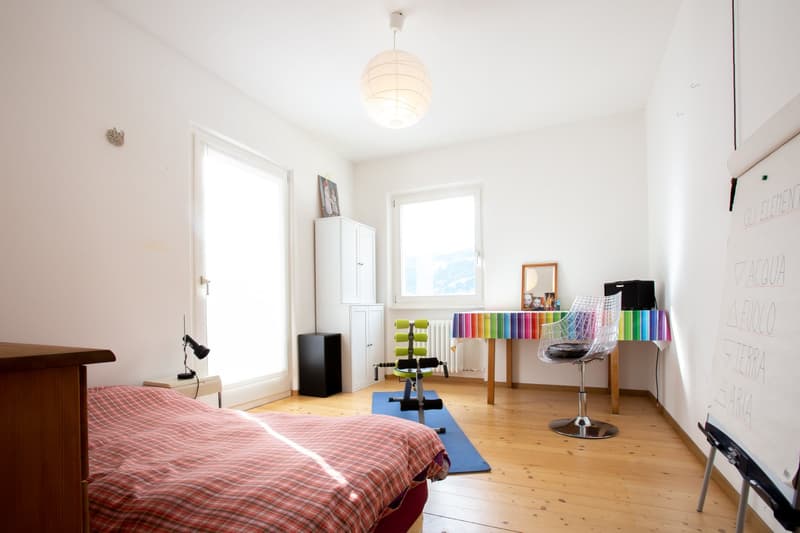 Bright and spacious bedroom with hardwood floors, large windows, a plaid bedspread, a white wardrobe, and a colorful display of paint swatches and art supplies.