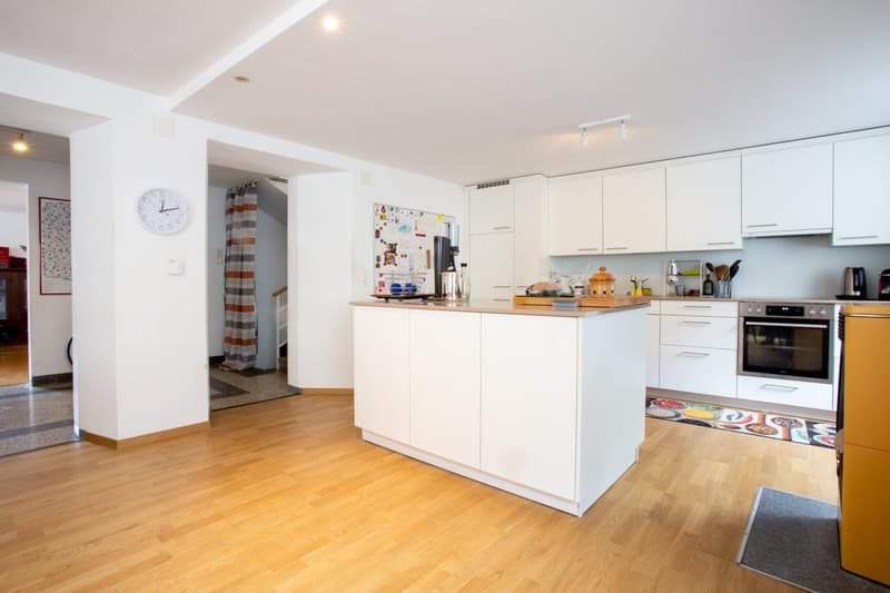 Spacious open-plan kitchen with white cabinets, wooden countertops, and hardwood flooring. The kitchen is equipped with a dishwasher and various kitchen appliances. There is a clock on the wall and a colorful rug on the floor.