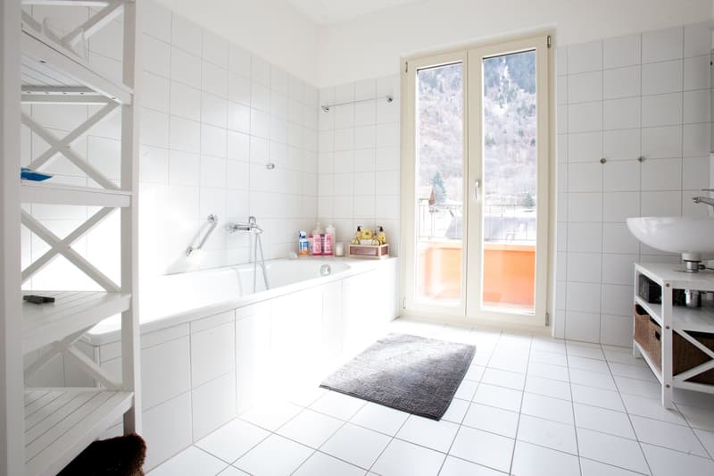 Large bathroom with white tiled walls and floor, bathtub, shelves with toiletries, and a window overlooking the outdoors.
