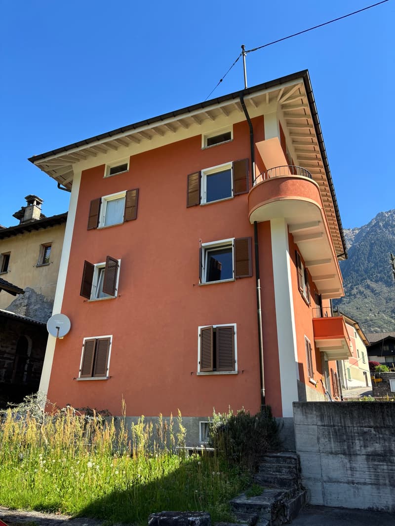 2-story residential building with orange exterior walls, wooden shutters, and a balcony on the upper floor. The building is situated in a mountainous area with a grassy area in the foreground.