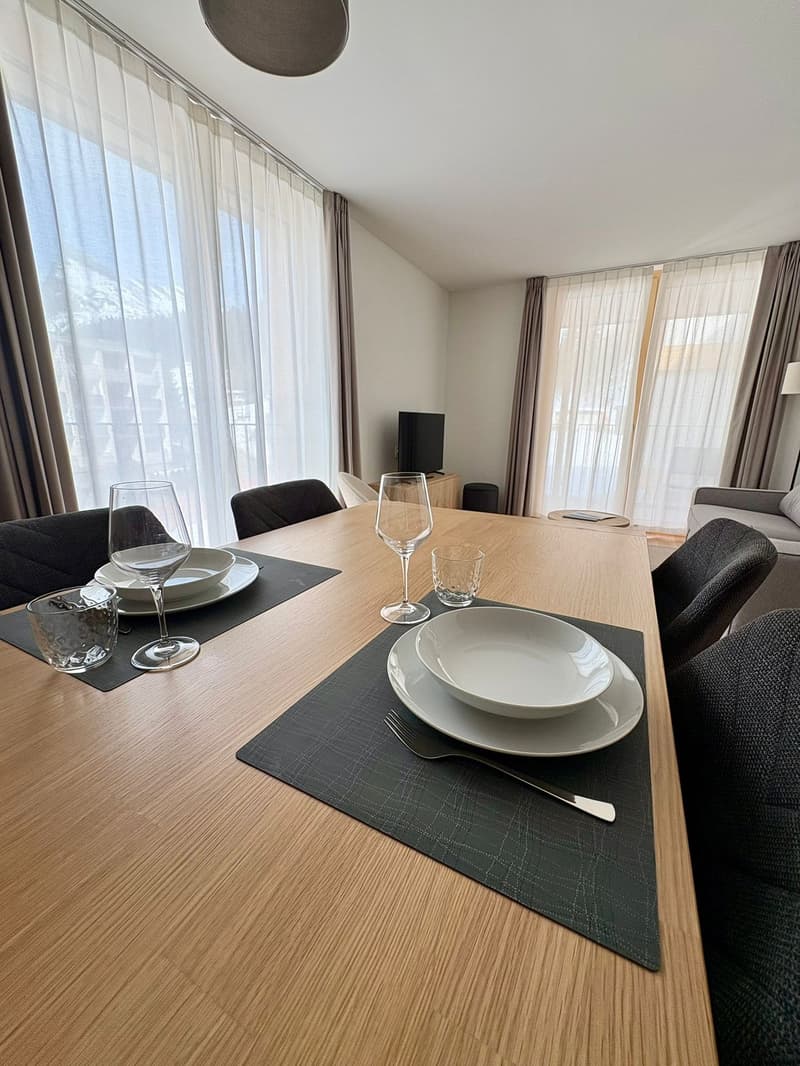 Dining area with a wooden table, wine glasses, plates, fork, spoon, a television, and a window with curtains