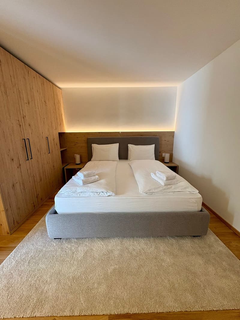Spacious bedroom with double bed, wooden closets, bedside tables, white walls, and a rug.