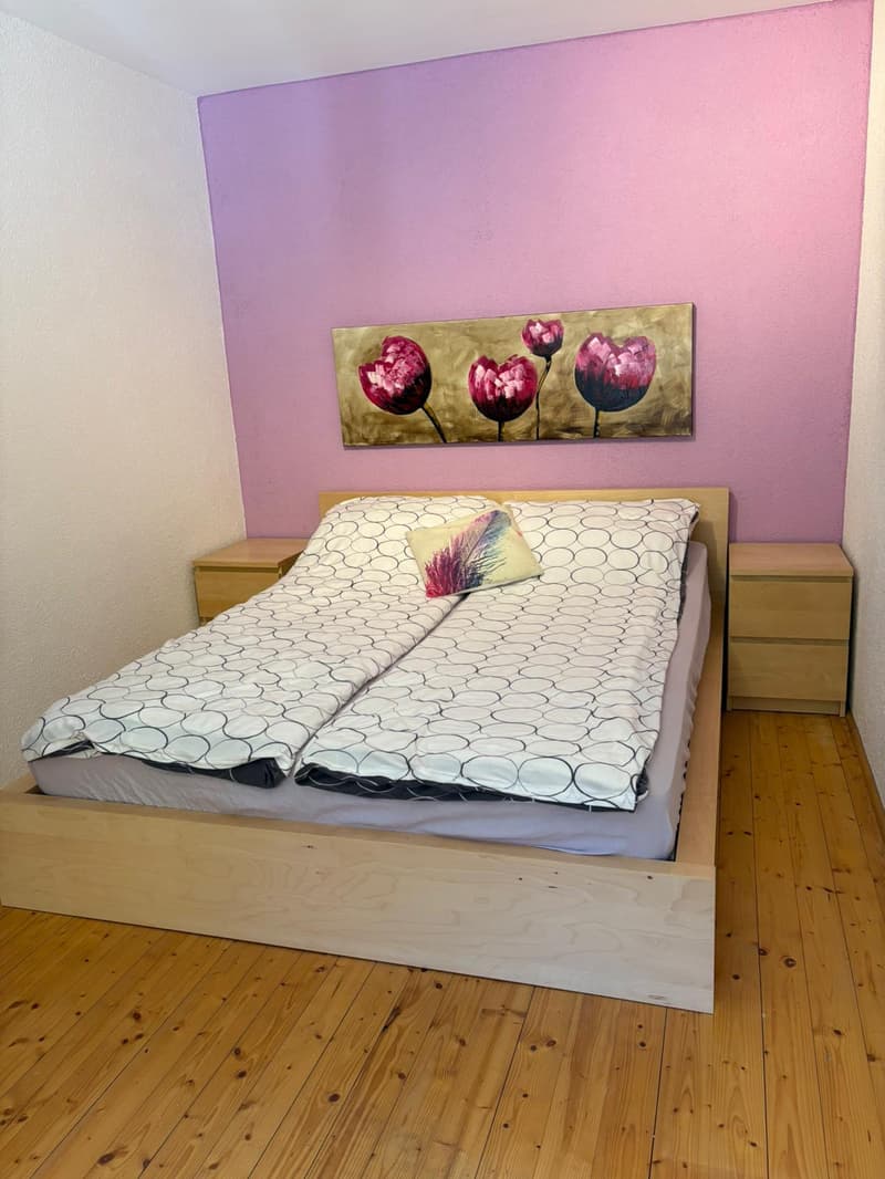 Bedroom with two single beds, wooden floor, pink wall, wooden nightstands, decorative pillows, and a large painting above the bed.