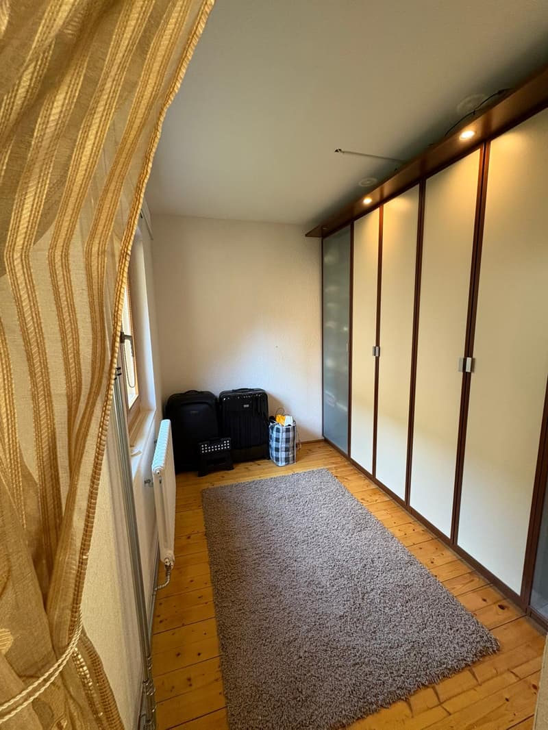 Empty room with wooden floor, white walls, large wardrobe, rug, luggage, and a window with a curtain