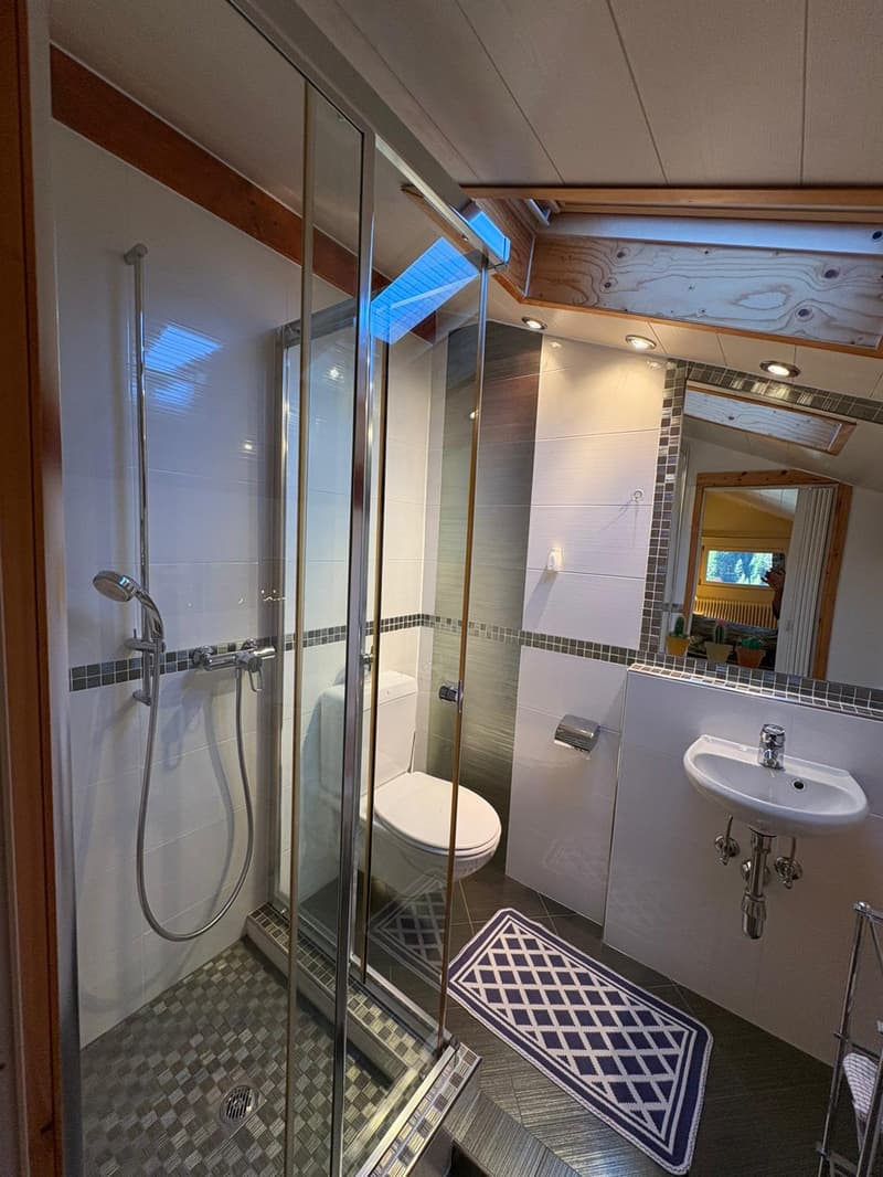 modern bathroom with glass shower, white toilet, white sink, mirror, wooden floor, tiled walls