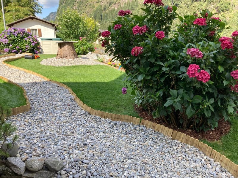A garden with a gravel pathway, green lawn, and various plants and flowers. There is a wooden table and a house in the background.