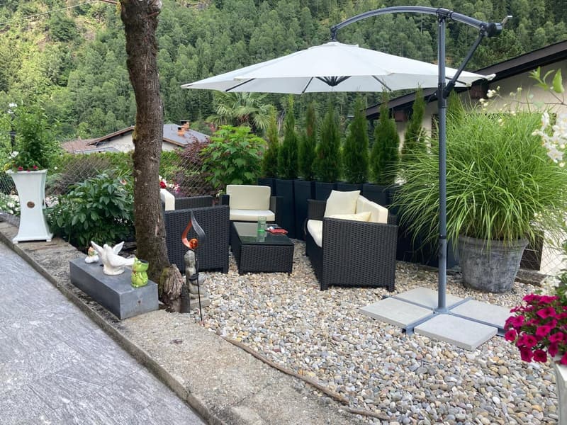 Outdoor seating area with a table, chairs, and an umbrella, surrounded by plants and flowers, with a scenic mountain view in the background.