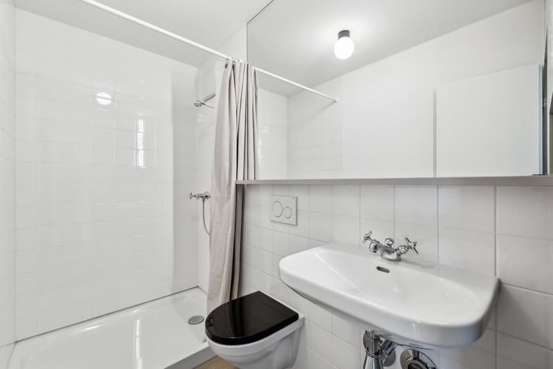 The image shows a modern, minimalist bathroom with white tiles, a pedestal sink, a toilet, and a shower curtain. The room has a clean and simple design with a single light fixture on the ceiling.