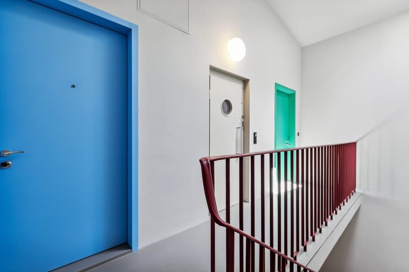 Bright and modern hallway with a blue door, a white wall, and a red railing leading to other rooms with colored doors