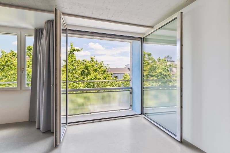 Spacious room with large windows and sliding glass doors leading to a balcony or terrace. The room has a concrete ceiling and gray curtains. The view outside shows lush greenery and a cloudy sky.