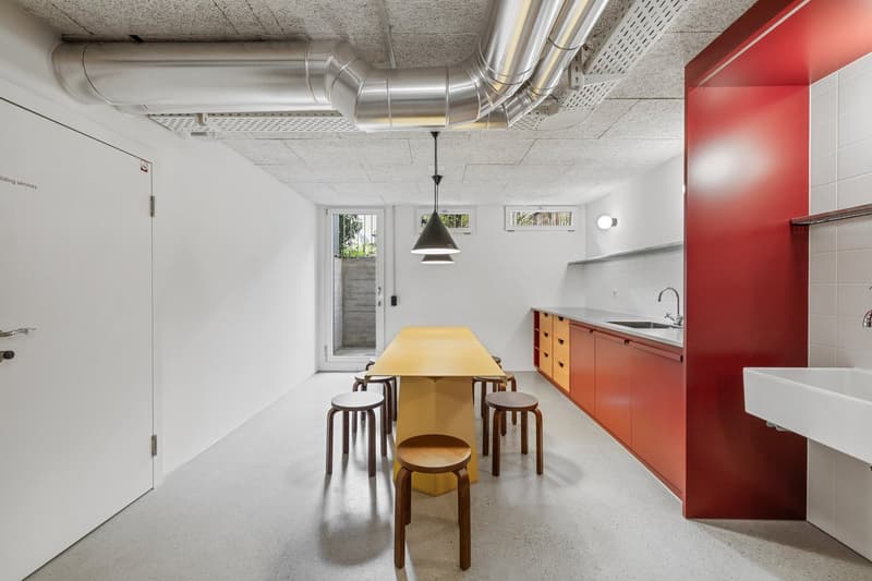 The image shows a modern, open-plan kitchen and dining area. The kitchen features a red cabinet unit with a sink and what appears to be a dishwasher. The dining area has a large wooden table with stools, and the space is illuminated by a pendant light. Th